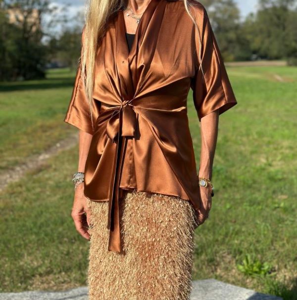 Donna indossa un top in raso color bronzo annodato in vita e una gonna con frange dorate, in un look sofisticato e glamour ambientato all’aperto.