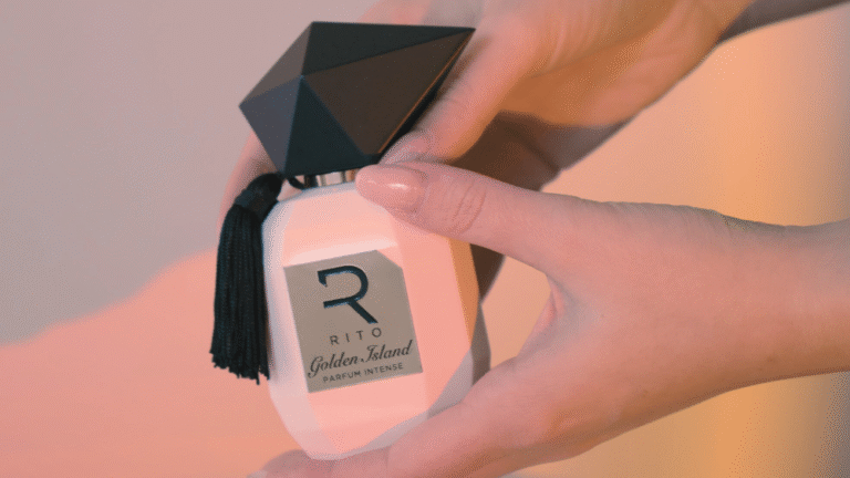 Mani femminili che tengono il flacone del profumo RITO Golden Island, illuminato da una luce calda.
