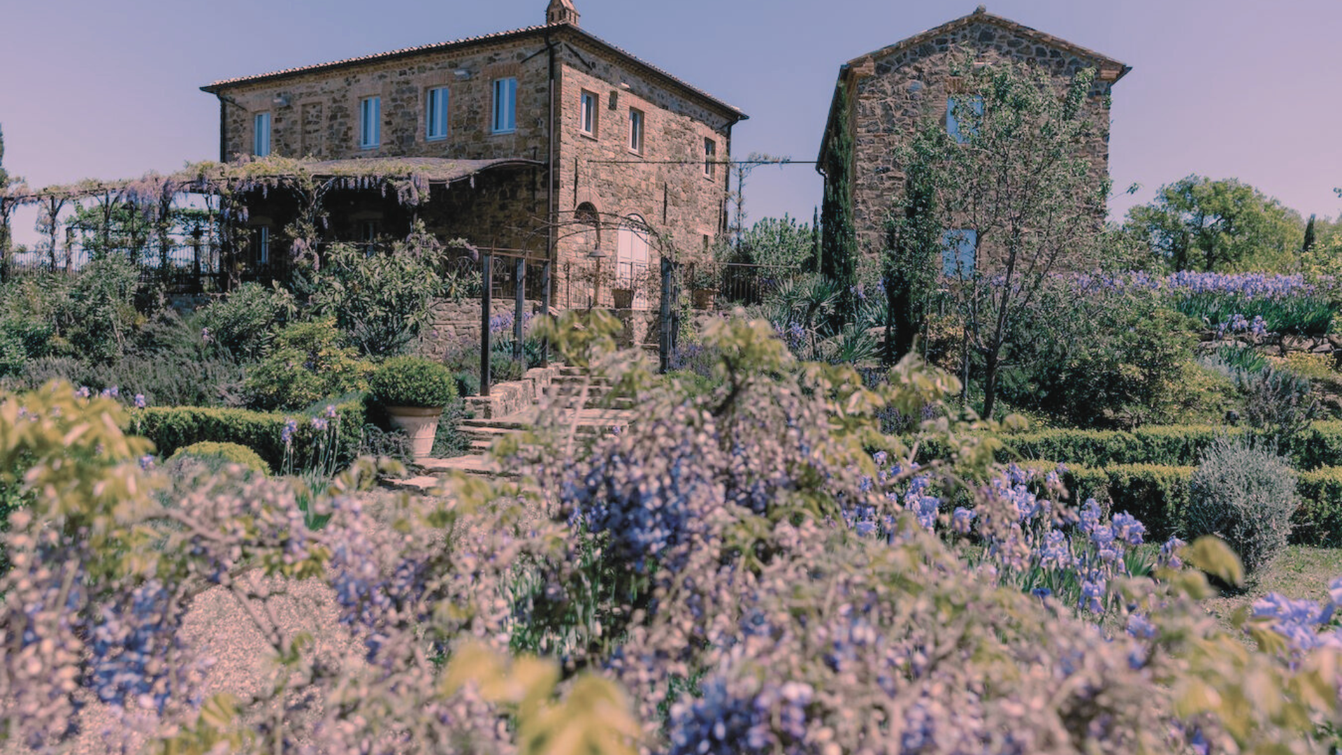 Casale in pietra della Tenuta Luce a Montalcino circondato da un rigoglioso giardino fiorito con glicini e iris in fiore, immerso nella campagna toscana.
