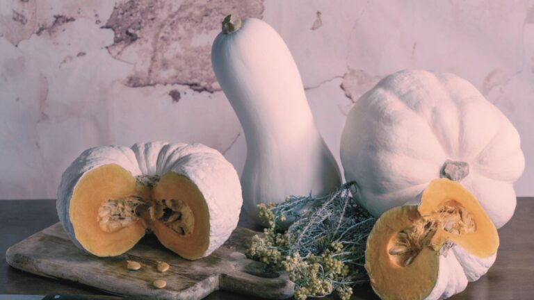 Zucche autunnali intere e tagliate a metà disposte su tagliere di legno con fiori secchi, in una composizione rustica e naturale.