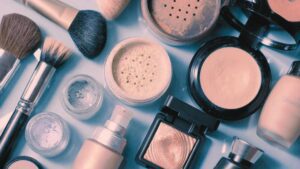 Flat lay di cosmetici e accessori make-up su fondo azzurro: pennelli, ciprie compatte e in polvere libera, fondotinta con dispenser, ombretti e flacone di smalto.