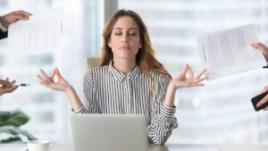 Donna seduta alla scrivania con laptop, occhi chiusi in atteggiamento meditativo mentre mani esterne le porgono documenti e oggetti.