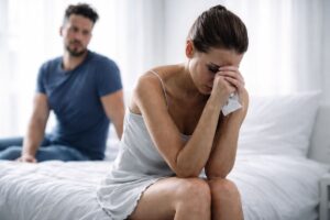 Donna seduta sul letto visibilmente provata mentre l’uomo resta sullo sfondo durante un momento di difficoltà nella coppia