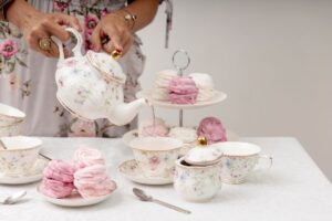 Donna versa il tè da una teiera in porcellana decorata con motivi floreali su una tavola apparecchiata per un tea party, con tazze coordinate e meringhe rosa e bianche disposte su alzate e piattini.