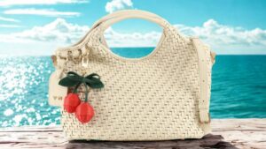 Borsa intrecciata color avorio con charm a forma di ciliegie, fotografata davanti a un mare turchese.