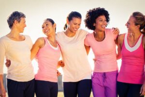 Cinque donne sorridenti, vestite con abiti sportivi nei toni del rosa, si abbracciano all’aperto in un’atmosfera luminosa e positiva.