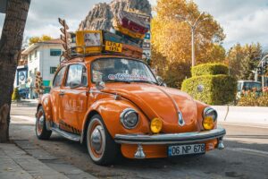 Maggiolino arancione d’epoca parcheggiato in strada, con valigie e bagagli fissati sul portapacchi.