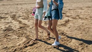 Due ragazze camminano sulla sabbia indossando bermuda estivi, con pattini e accessori tra le mani.