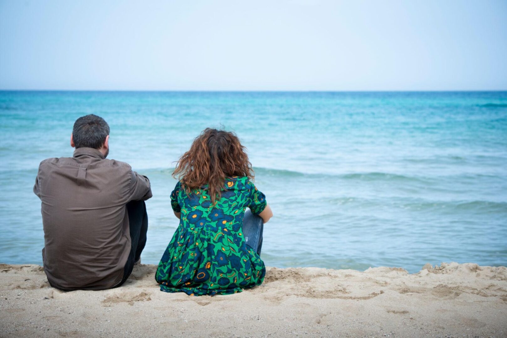 Coppia seduta sulla spiaggia, di spalle e a distanza, davanti al mare, in un momento di silenzio e tensione emotiva.