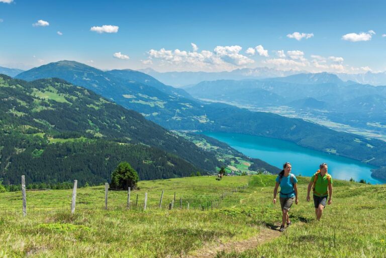 Due escursionisti percorrono un sentiero tra i prati verdi della Carinzia, in Austria, con vista panoramica su un lago dalle acque turchesi e sulle montagne alpine.