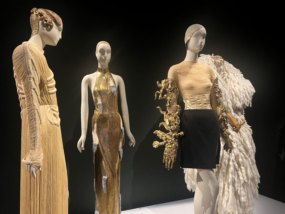 Manichini con abiti dalle forme surrealiste: un vestito beige con rilievi anatomici, un abito lungo dorato e un completo con corsetto chiaro, gonna nera e decorazioni dorate sulle braccia.