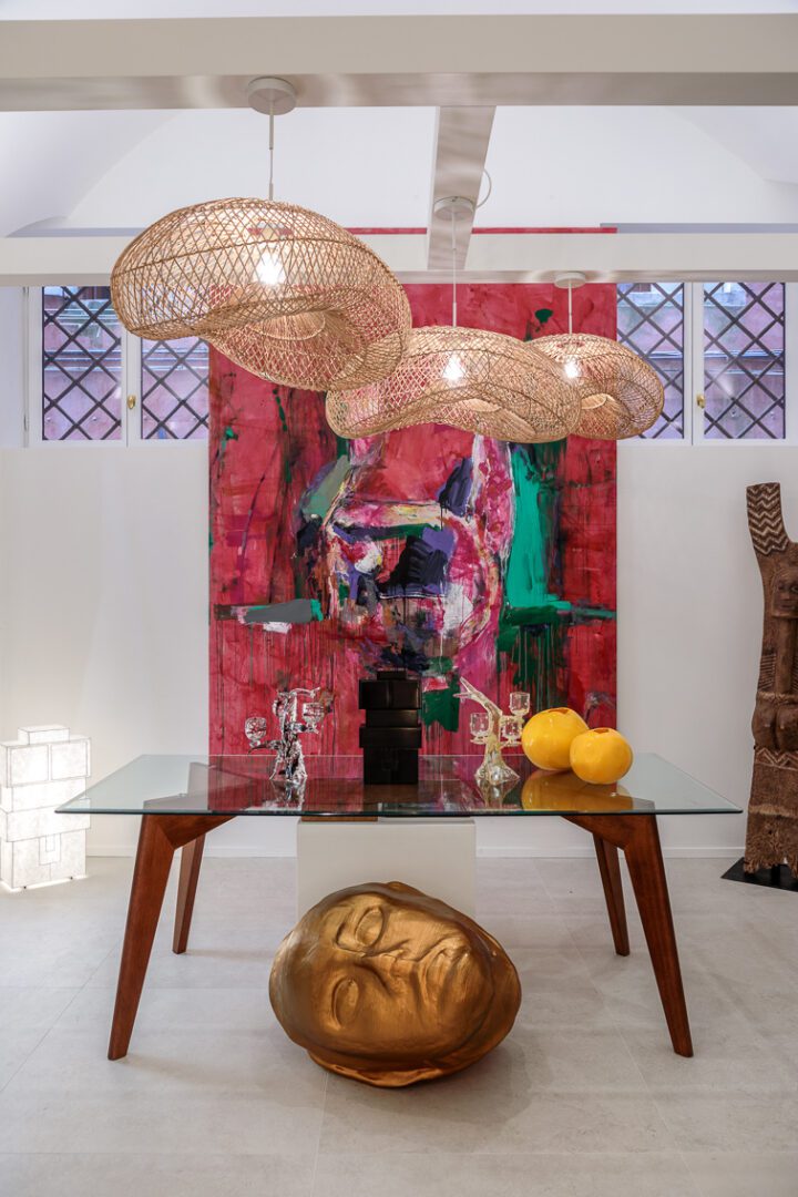 Interno de La Bottega della Fondation Valmont a Venezia, con lampade sospese intrecciate, un grande dipinto astratto e una scultura dorata raffigurante un volto.