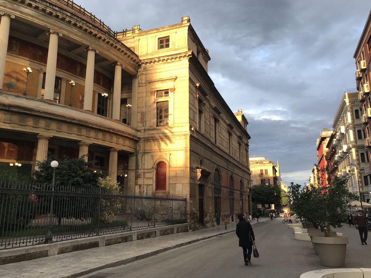 Teatro Politeama Garibaldi di Palermo illuminato dalla luce dorata del tramonto, con il suo colonnato monumentale e la strada pedonale alberata.
