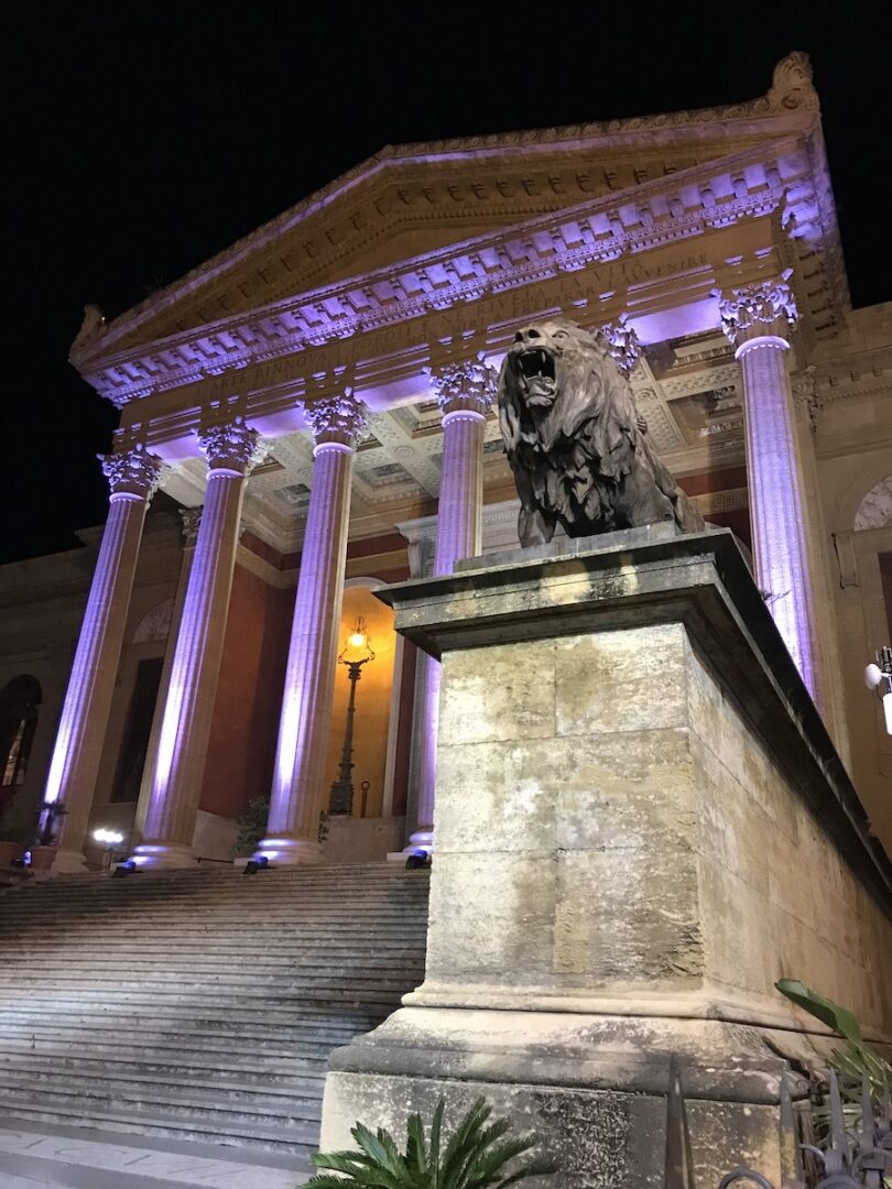Il Teatro Massimo di Palermo illuminato di viola durante la sera, con la monumentale scalinata, le colonne corinzie e una delle sculture raffiguranti un leone in primo piano.