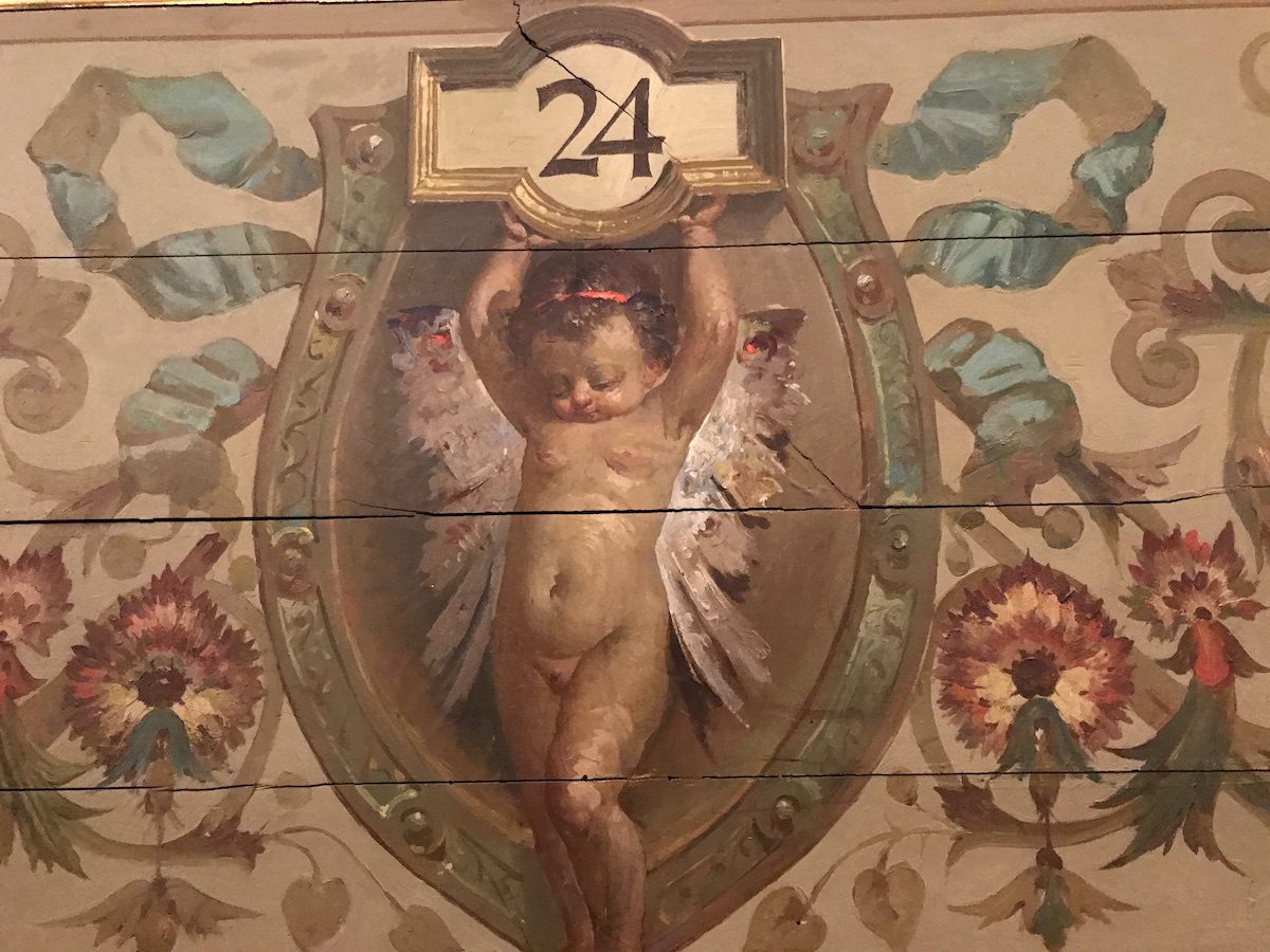 Dettaglio delle decorazioni pittoriche del Teatro Massimo di Palermo, con un putto alato che sorregge il numero 24, circondato da motivi floreali e ornamentali.