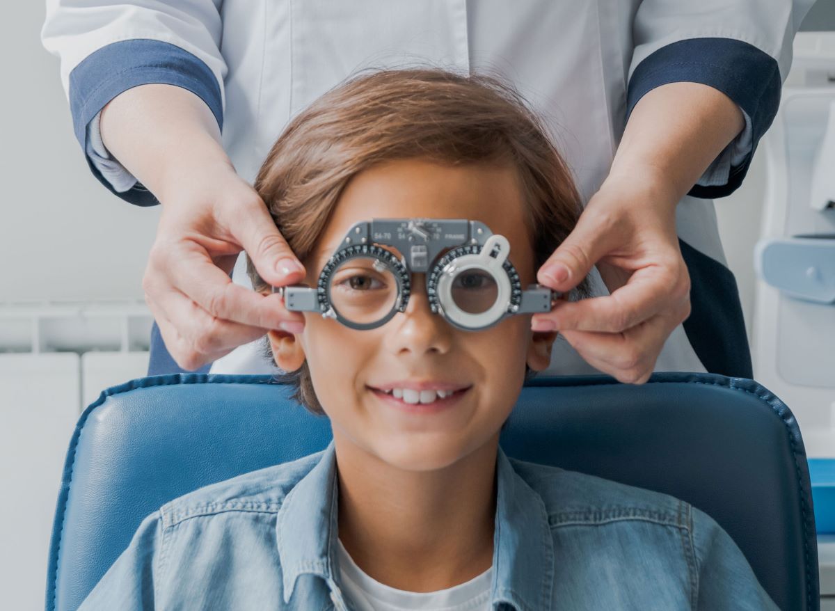 Bambino durante una visita oculistica, mentre lo specialista gli posiziona una montatura di prova con lenti graduate per valutare eventuali difetti visivi, come la miopia infantile.