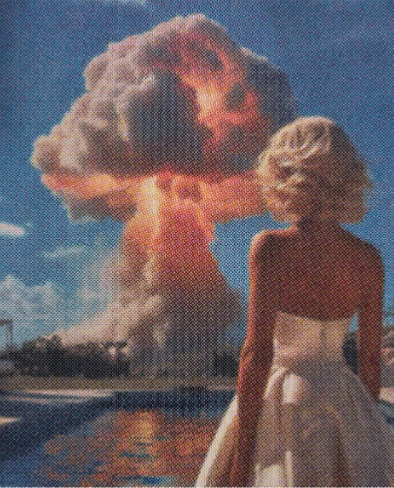Donna bionda di spalle, con abito bianco, davanti a un'enorme nube a fungo illuminata dai bagliori di un'esplosione